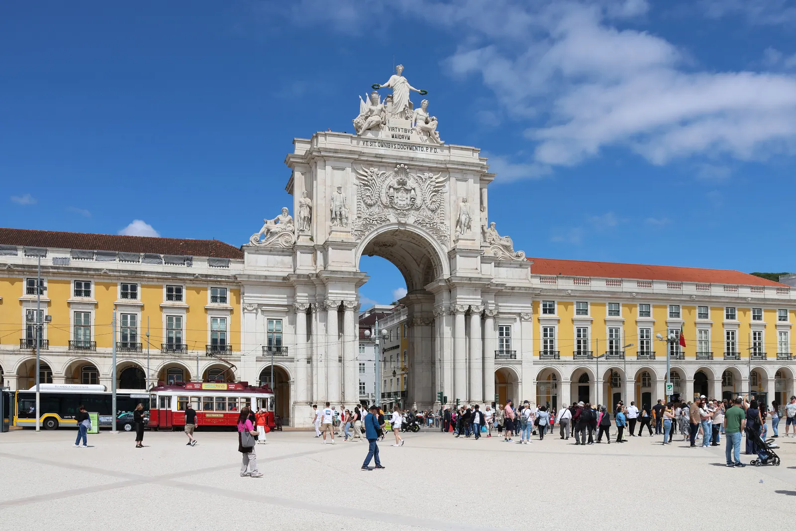 The triumphal Rua Augusta Arch between the yellow arcades of Praça do Comércio in Lisbon, with a red tram and strolling visitors under a blue sky