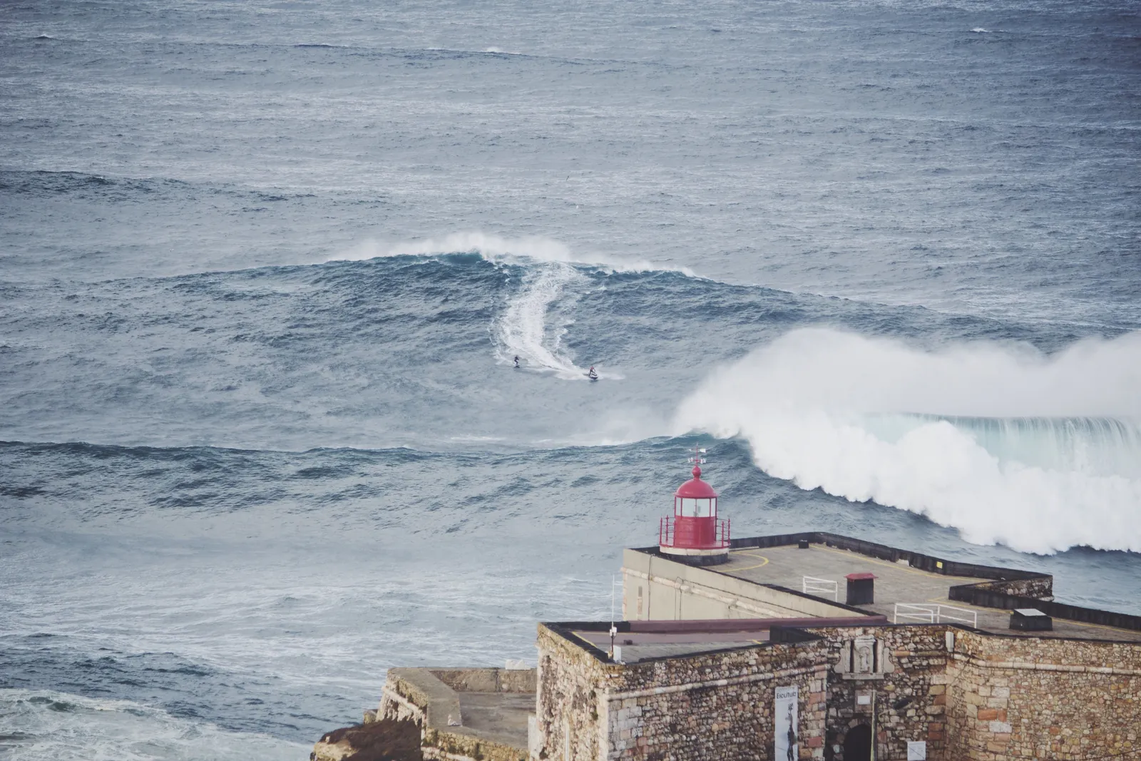 A tow-in surfer riding a giant Praia do Norte wave beyond the red lighthouse of the Nazaré fort