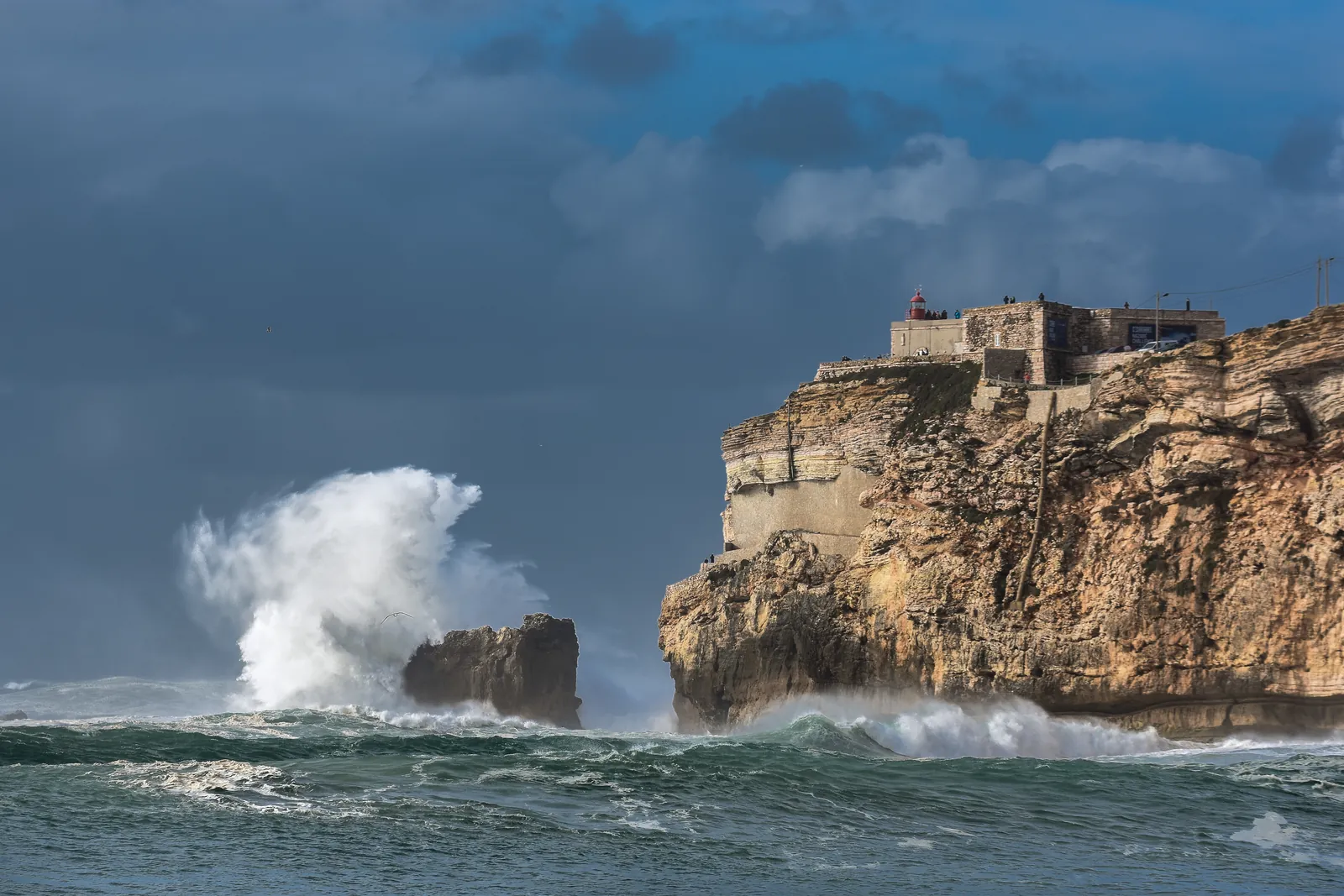 Waves breaking below the São Miguel Arcanjo fort and its red lighthouse on the Nazaré headland