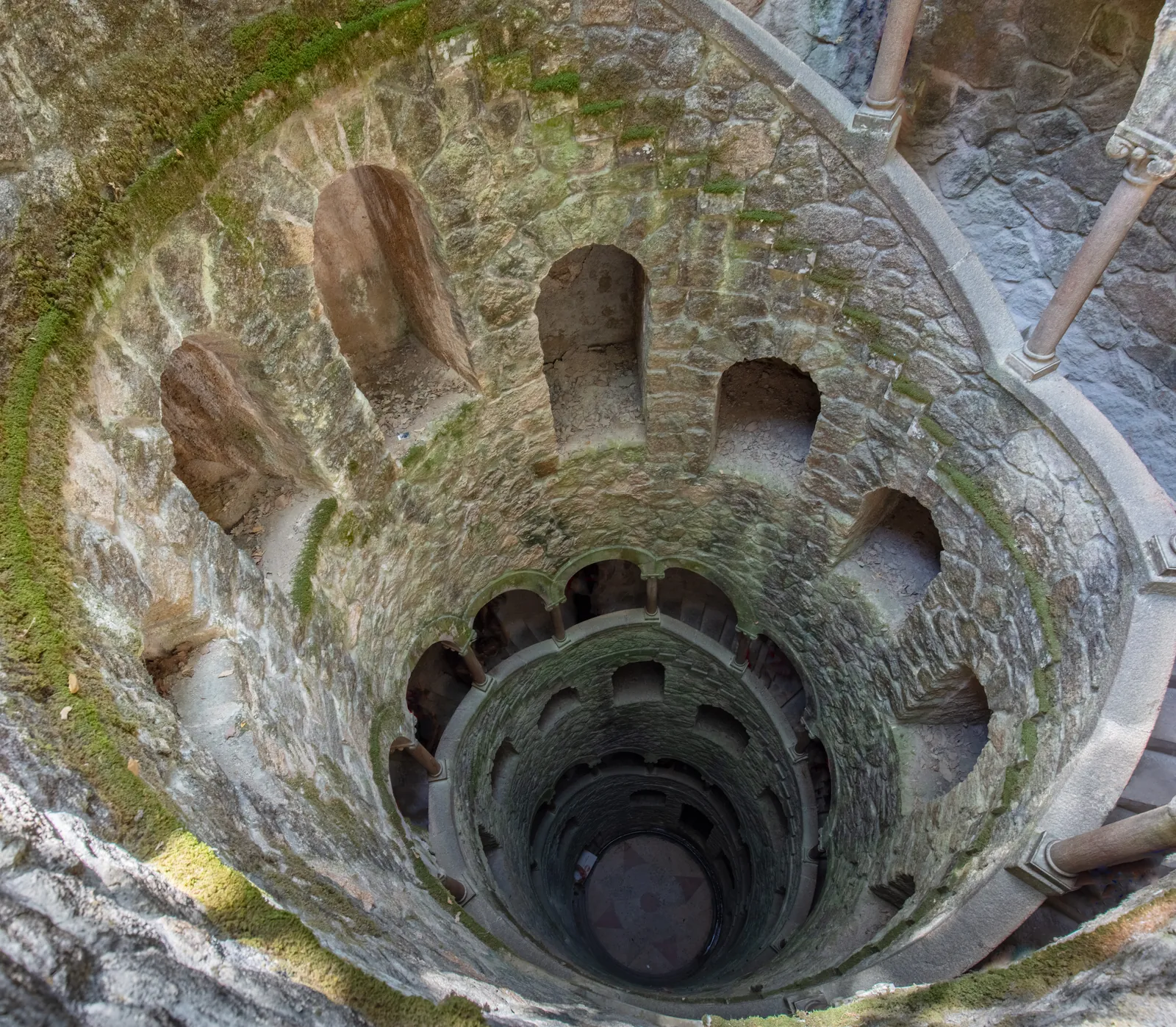 Looking down the mossy spiral of the Initiation Well at Quinta da Regaleira in Sintra, its arcaded stone galleries descending into shadow